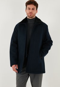 Námořnický modrý kabát s černým měkkým límcem, předním zipem a bočními kapsami, vyrobený z tlusté, strukturované tkaniny. Model pózuje s rukama v kapsách.