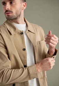 Tan jakke med krave, sorte knapper og en brystlomme, båret over en hvid skjorte. Glat stof med en afslappet pasform, der fremhæver armene.