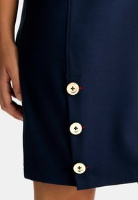 Abito blu navy realizzato in tessuto morbido, caratterizzato da quattro grandi bottoni laterali con dettagli rossi e un taglio dritto elegante.