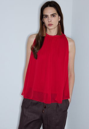 Kobieta o długich brązowych włosach nosi czerwoną bluzkę bez rękawów, brązowe spodnie i długie kolczyki z frędzlami, stoi na tle gładkiej jasnej ściany.