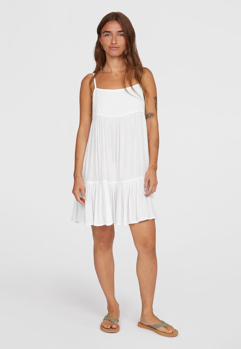 Weißes ärmelloses Kleid mit quadratischem Ausschnitt, strukturiertem Stoff und einem mehrlagigen Rüschensaum. Kombiniert mit braunen Flip-Flops.
