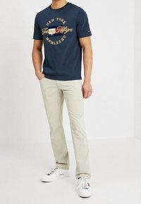 Camiseta de manga corta azul marino con el logotipo de "Tommy Hilfiger" y acentos dorados, combinada con unos vaqueros beige claros y zapatillas blancas.