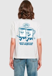 Weißes T-Shirt mit blauer Grafik von zwei Kaffeetassen mit Gesichtern, der Text lautet "Trink deinen täglichen Espresso" und "mehr Espresso, weniger Depressio."