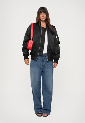 Black bomber jacket with ribbed cuffs, white T-shirt, blue wide-leg jeans, and a red crossbody bag with a circular logo. Flat sandals.