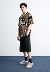 Camo trøje med nummer "03," sorte akcenter og Adidas-logo; parret med sorte knælange shorts og grå sneakers med røde striber.