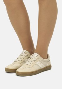 Zapatillas beige con parte superior de cuero suave, puntera redondeada, diseño plano con cordones, y suela de goma texturizada. Logo visible en la lengüeta.