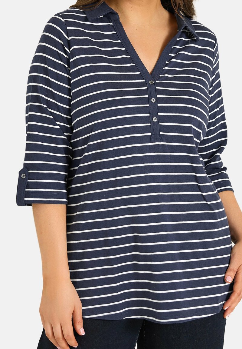 Femme portant une chemise à manches longues rayée bleu marine et blanche avec col boutonné et manches retroussées, associée à un pantalon foncé.