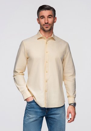 Mand med kort mørkt hår iført en cremefarvet skjorte med knapper og blå jeans, stående med den ene hånd i lommen mod en ensfarvet baggrund.