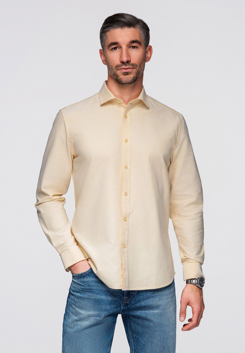 Homme aux cheveux courts foncés portant une chemise crème boutonnée et un jean bleu, debout avec une main dans la poche devant un fond uni.