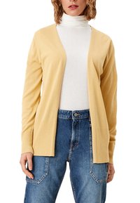 Żółty, prążkowany kardigan z otwartym przodem, z dwoma przednimi kieszeniami, noszony na białym golfie, w połączeniu z niebieskimi dżinsami z wysokim stanem.