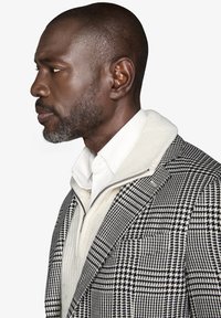Giacca da uomo in tessuto a scacchi bianco e nero, realizzata in misto lana, con revers a picco, indossata sopra un maglione zip-up beige con colletto.