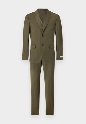 Muž v olivovo zelenom obleku so svetlo béžovým pleteným polotrickom a čiernymi spoločenskými topánkami, stojaci s rukami v vreckách na jednofarebnom pozadí.