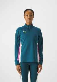 Türkiser Sporttop mit Reißverschluss und pinken sowie weißen Seitenpaneelen. Verfügt über einen hohen Kragen und lange Ärmel; hergestellt aus einem glatten, dehnbaren Stoff.