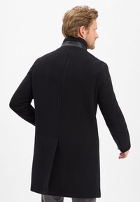 Schwarzer Mantel mit hohem Kragen aus strukturiertem Stoff, mit geteiltem Rücken, langen Ärmeln und kontrastierenden Lederakzenten am Kragen.