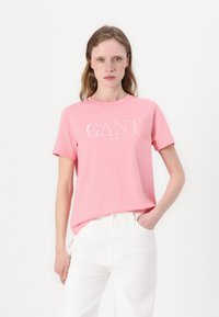 Rosa bomullströja med rundad halsringning och korta ärmar, med "GANT"-logotypen tryckt i vitt på framsidan. Parat med vita byxor.