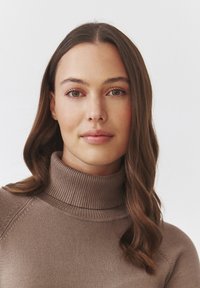 Brązowy sweter z golfem o prążkowanej fakturze, dopasowanym kroju i bezszwowych ramionach. Modelka ma długie, falujące włosy i naturalny makijaż.