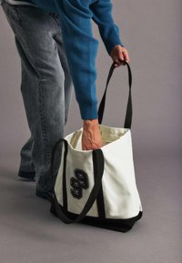 Bolsa de lona en blanco con una base marrón oscuro. Presenta el número negro "99" en el lateral y dobles asas negras. La textura parece resistente.