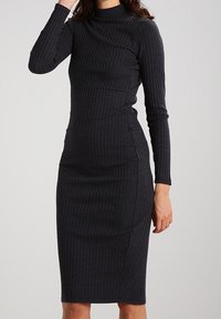 Svart ribbad stickad klänning med långa ärmar och hög halsringning, figurnära siluett och knälång nederkant. Texturerad tygdetalj synlig.