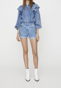 Femme portant un chemisier en denim bleu clair avec des épaules à volants, un short en denim assorti retroussé et des bottines blanches, sur fond blanc.