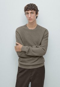 Brauner, strukturiert gestrickter Pullover mit Rundhalsausschnitt, kombiniert mit dunkelbraunen Hosen. Das Model steht mit verschränkten Armen vor hellem Hintergrund.