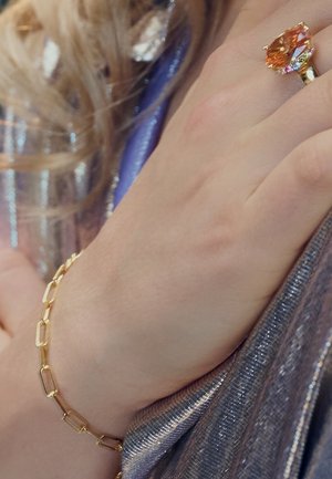 Guld kædearmbånd med rektangulære led, båret på en hånd, med en stor orange ædelsten ring. Baggrunden viser skinnende stof.