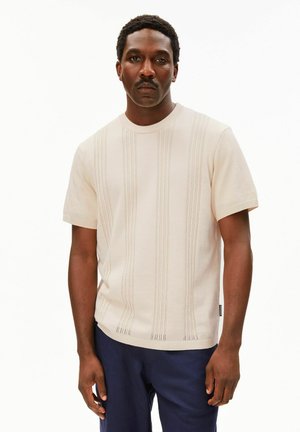 Homme aux cheveux courts et bouclés avec une moustache, portant une chemise beige à manches courtes avec des motifs tricotés verticaux et un pantalon bleu foncé, debout.