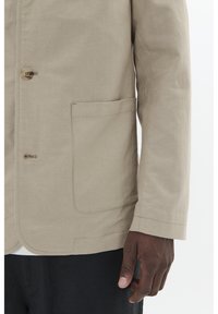 Light beige linen jacket featuring a single button, a front pocket, and a straight hem. The texture is smooth with visible stitching details.