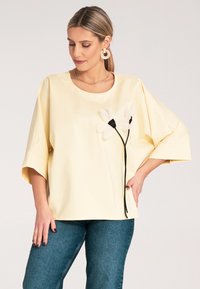 Żółta oversized bluzka z okrągłym dekoltem, krótkimi rękawami oraz czarnymi haftowanymi kwiatami z przodu. Połączona z niebieskimi dżinsami.