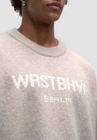 Beżowy sweter dziany z żeberkowym okrągłym dekoltem, z białym napisem "WRSTBHVR" i "BERLIN" wyraźnie widocznym na przodzie.