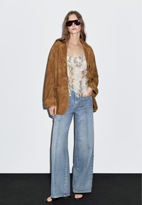 Brun suedetop oversized jakke, cremet slangeprint top med flæser, lyseblå brede jeans og mørke solbriller. Glatte teksturer.
