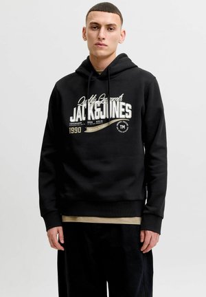 Młody mężczyzna w czarnej bluzie z kapturem z białym napisem „JACK & JONES” i beżową podkoszulką, stojący na tle jednolitego jasnego tła.