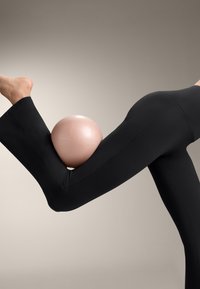 Čierne legíny s hladkou textúrou, natiahnuté cez ohnutú nohu, drží svetloružovú loptu medzi stehnom a lýtkom.