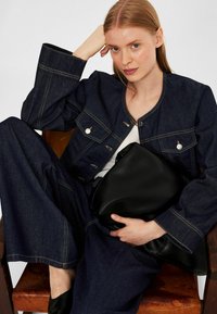 Kvinna med ljus hy och röd hår sitter på en brun stol, klädd i en mörk denimjacka och byxor, håller en stor svart läderhandväska.