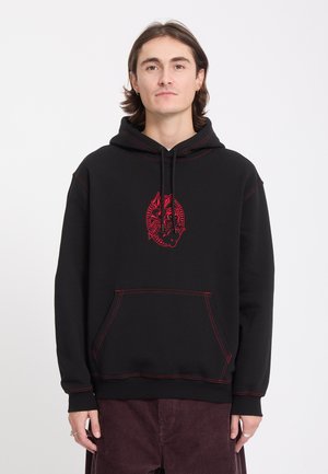 Jeune homme aux cheveux mi-longs portant un sweat à capuche noir avec un motif de dragon rouge et des coutures rouges assorties, debout devant un fond blanc.