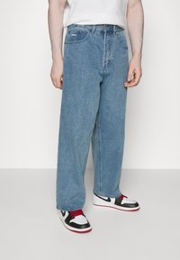 Light blue denim jeans with a loose fit, five-pocket design, and logo detail on the waistband. Paired with black and white sneakers.