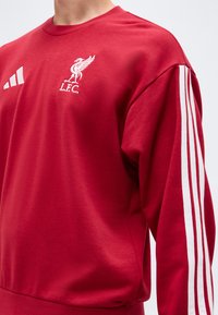 Κόκκινο φούτερ με λευκό έμβλημα της Liverpool FC και λογότυπο Adidas στο στήθος, με τρεις λευκές ρίγες στα μανίκια.