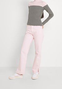 Henkilö, joka on pukeutunut harmaaseen ja vaaleanpunaiseen pitkähihaiseen neuleeseen, vaaleanpunaiseen housuun, joissa on nilkkaleikkaukset, ja valkoisiin lenkkareihin, seisoo yksinkertaista taustaa vasten.