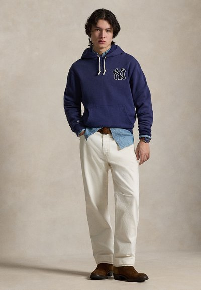 Marineblå hettegenser med hvit logo, snor, kombinert med lys denimskjorte, kremfargede bukser og brune støvler. Jevn stofftekstur overalt.