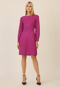 Strickkleid in leuchtendem Pink mit Zopfmuster im Oberteil, langen Ärmeln und gerade geschnittenem Rock. Getragen mit schwarzen Schuhen.