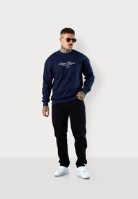 Navy blue sweatshirt with white logo, paired with black distressed jeans and white sneakers. Model also wears sunglasses.