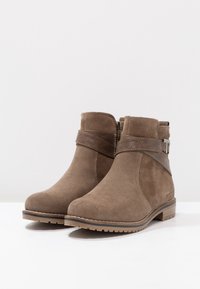 Paire de bottines en daim marron avec des sangles en cuir marron foncé et des boucles latérales, posées sur une surface blanche devant un fond uni.
