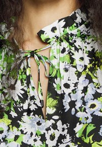 Floral blouse with a green, black, and white pattern. Features a gathered neckline and a tie closure at the front with black hardware accents.