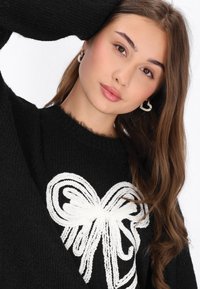 Czarny dziergany sweter z białym wzorem kokardy na przodzie. Modelka nosi okrągłe kolczyki, ma długie, faliste brązowe włosy.