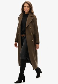 Bruine lange jas van een wollen mix met dubbelrijige knopen, grote revers, gecombineerd met een zwarte trui, donkere spijkerbroek en zwarte laarzen met hak.