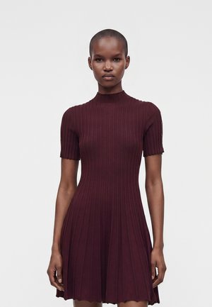 Femme portant une robe courte à manches courtes, côtelée, de couleur bordeaux foncé avec un col montant, debout sur un fond gris clair uni.