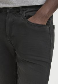 Melni denim džinsi ar gludu tekstūru, ar priekšējo kabatu un redzamu jostasvietu. Audums rāda nelielu nodilumu un ir pielāgots piemērs.
