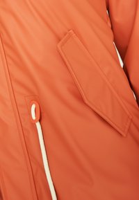 Veste imperméable orange avec une poche diagonale, un cordon en corde blanche et une texture lisse avec une finition mate.