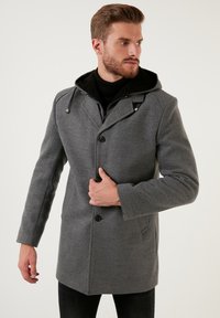 Abrigo de lana gris con capucha, cierre de botones doble y bolsillos laterales, con un acabado texturizado y acentos en negro.