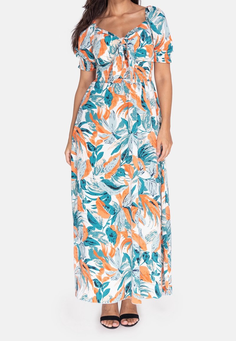 Femme portant une robe longue blanche avec des motifs floraux orange et turquoise, des manches bouffantes et des talons ouverts noirs.