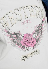 T-shirt bianco con testo grafico "WESTERN GIRL" e una rosa rosa con ali. Include un design a nastro con la scritta "SPIRIT OF THE WEST."